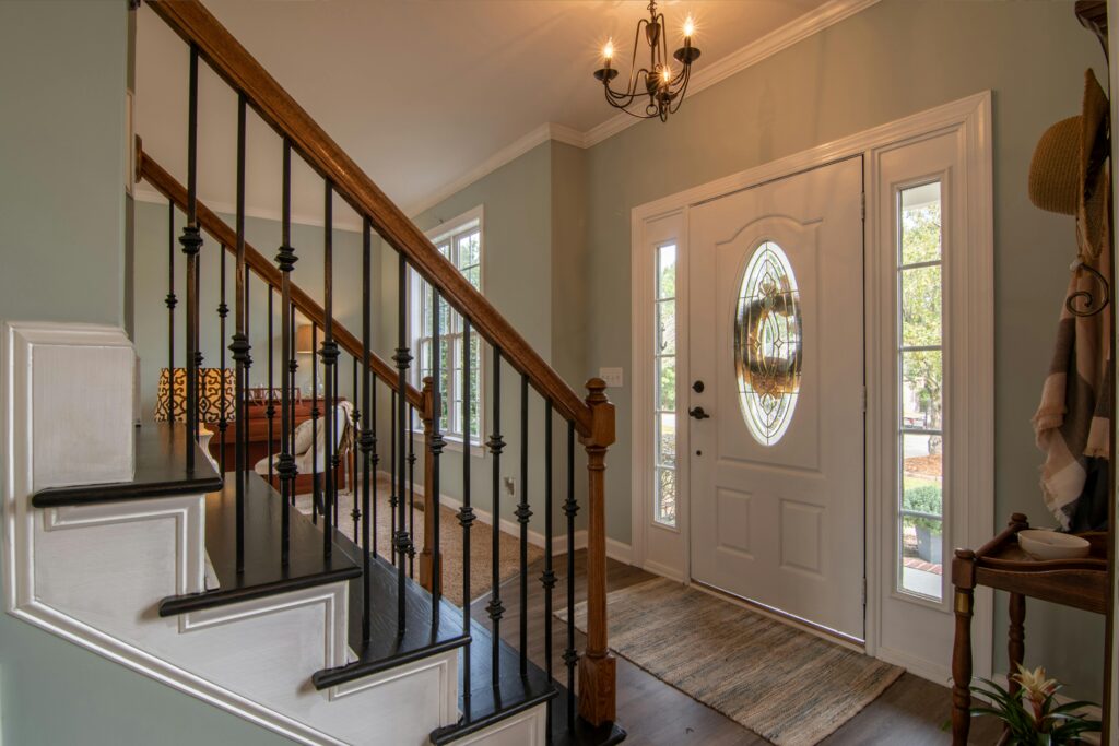 Elegant Entryway Of A Luxury Home Featuring A White Front Door With Decorative Glass, Hardwood Staircase With Wrought Iron Railings, And Bright Natural Light Showcasing The Spacious Interior Design.