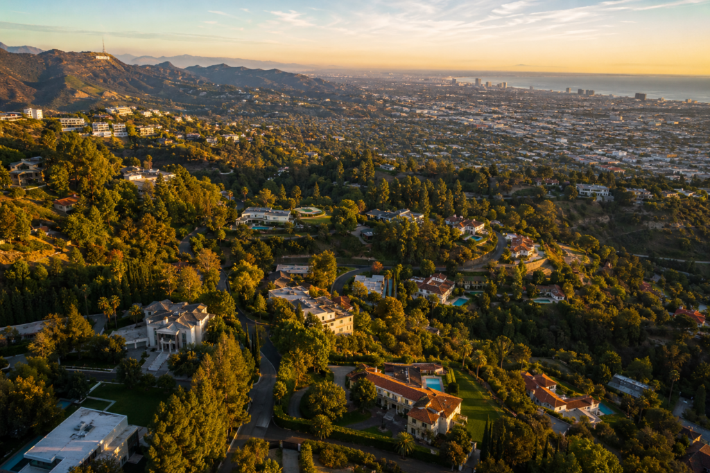 Aerial View Of Los Angeles Luxury Neighborhoods At Golden Hour With The Hollywood Sign, Sprawling Hillside Estates, Tree-Lined Streets, And The Pacific Ocean In The Distance.